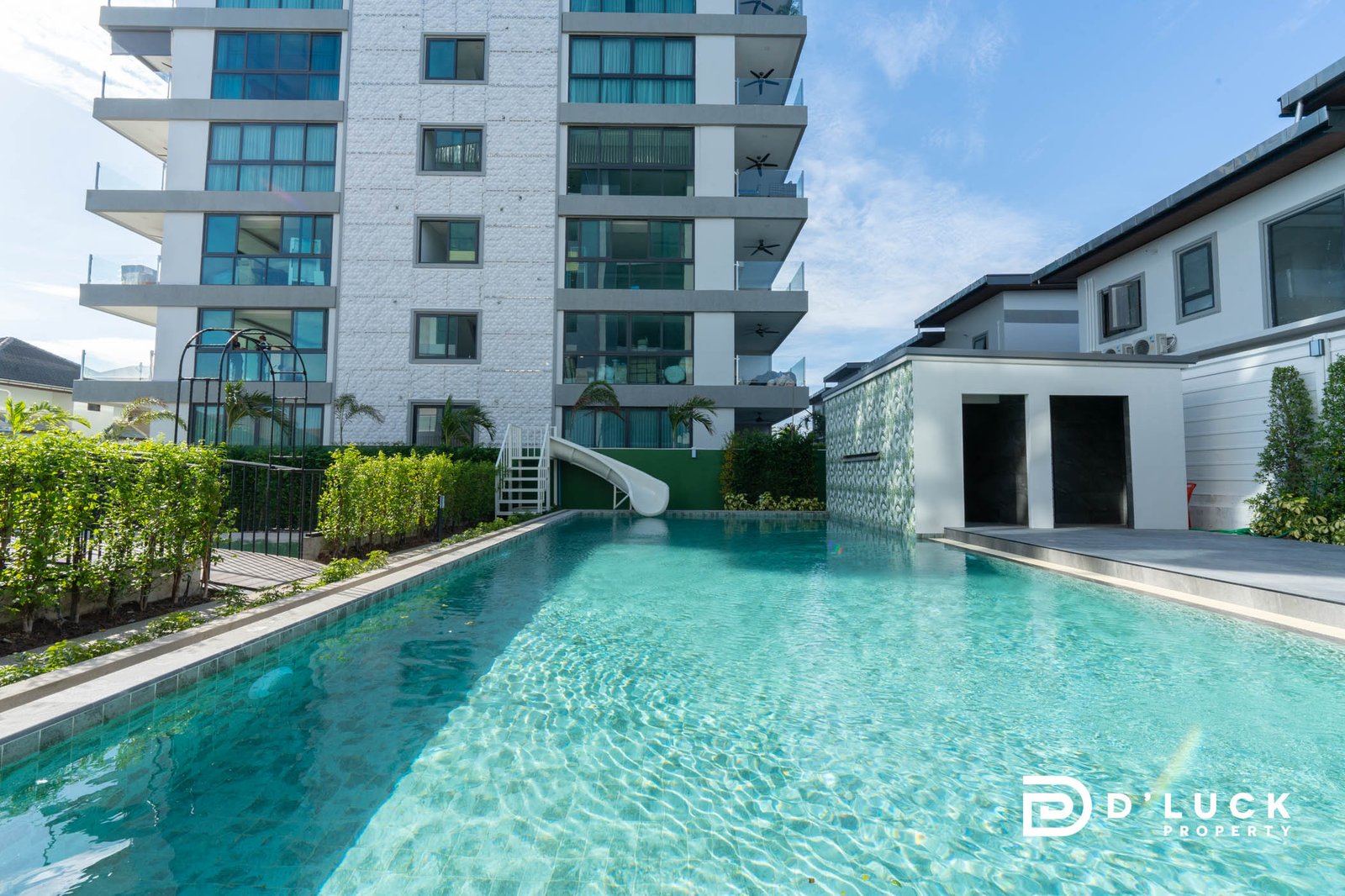Gardenia Pattaya 4 Modern white apartment building with glass balconies beside a long turquoise pool under a blue sky. Water slide and greenery at pool edge visible.