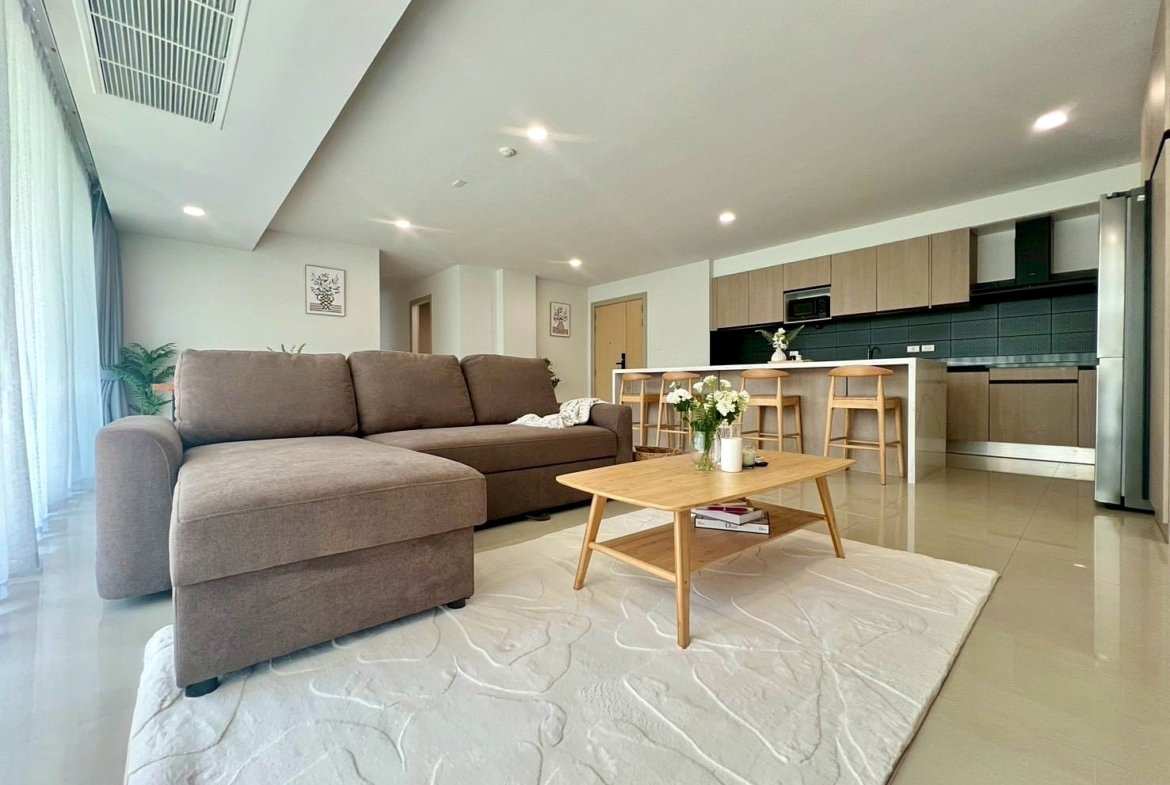 Open-concept living room with a brown sectional sofa, a wooden coffee table, and a bright kitchen island with four stools in the background.