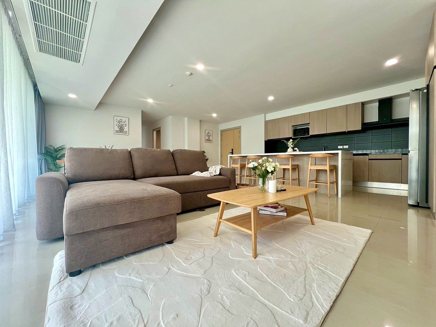 Gardenia_Pattaya_4Bed_Dlc492 Open-concept living room with a brown sectional sofa, a wooden coffee table, and a bright kitchen island with four stools in the background.