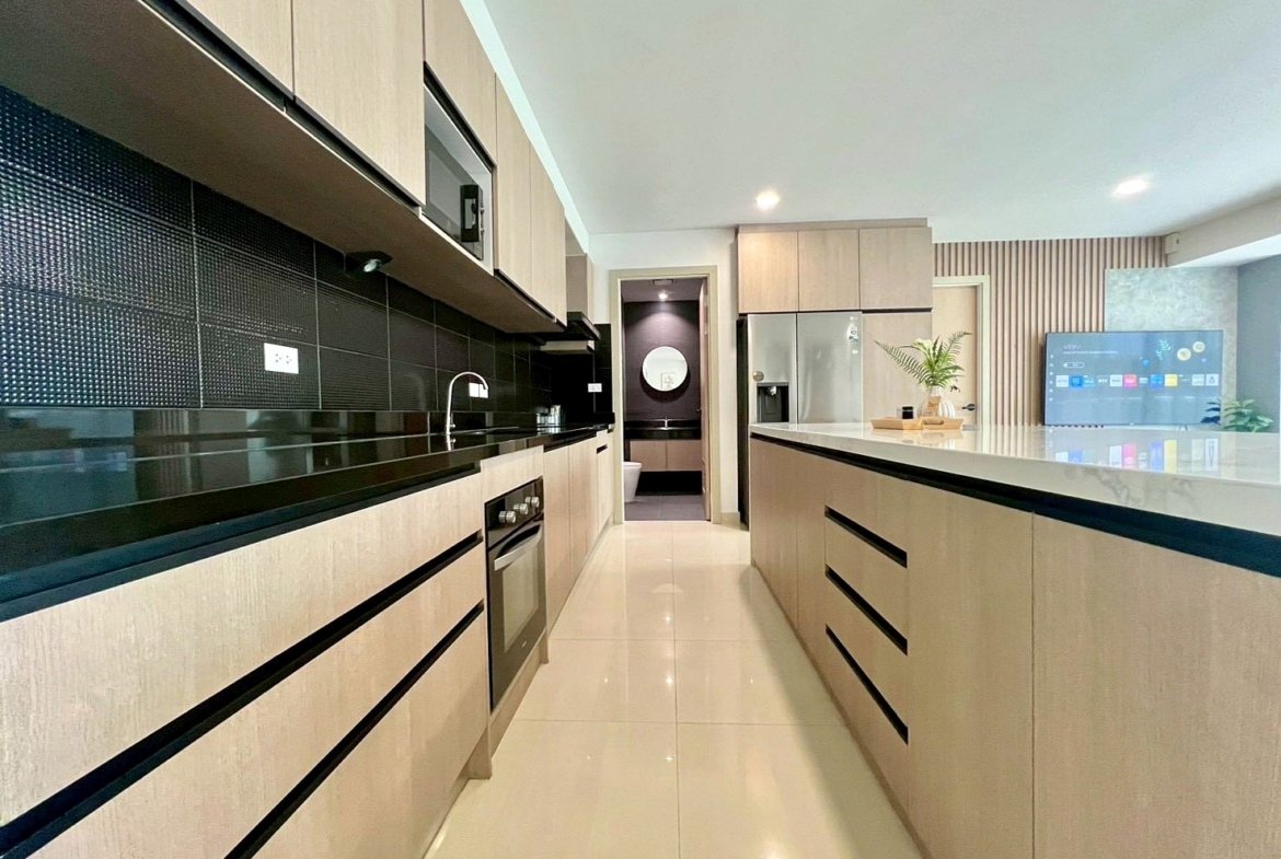 Modern kitchen with light wood cabinets, black backsplash, and a long island with a glossy white countertop; stainless steel appliances visible in the background.