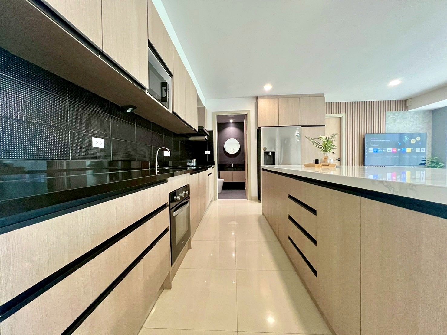 Gardenia_Pattaya_4Bed_Dlc492_12 Modern kitchen with light wood cabinets, black backsplash, and a long island with a glossy white countertop; stainless steel appliances visible in the background.