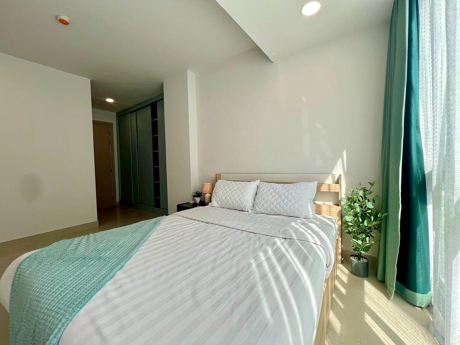 Gardenia_Pattaya_4Bed_Dlc492_13 Bright, minimalist bedroom with a wooden bed, white striped bedding, turquoise throw, and a potted plant by the window.