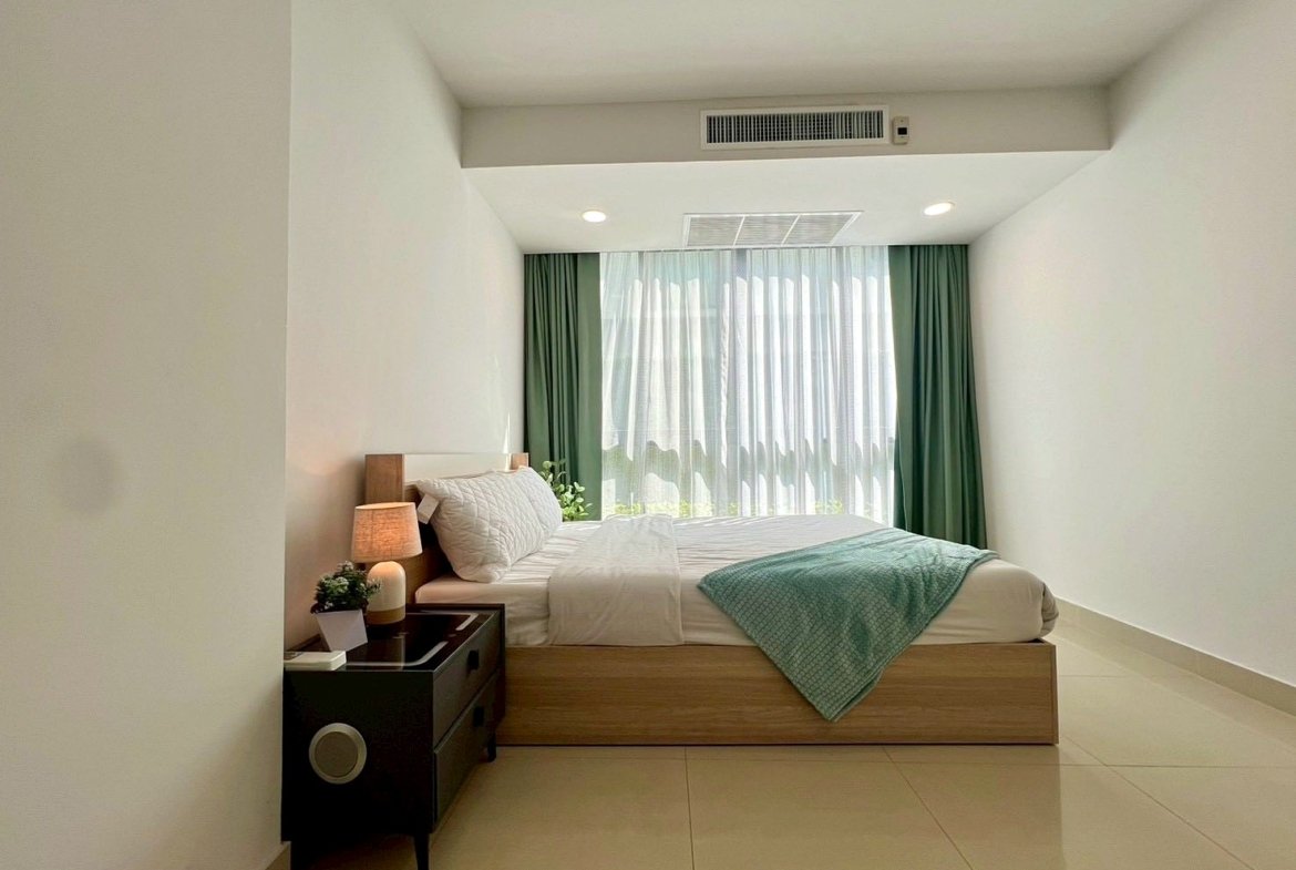 Bright hotel-style bedroom with a low wooden bed, white bedding, a turquoise throw, and a large window with sheer white curtains and green drapes.