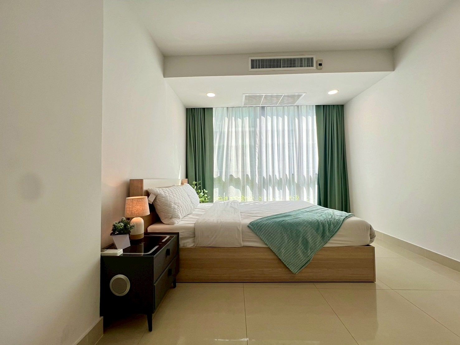 Gardenia_Pattaya_4Bed_Dlc492_16 Bright hotel-style bedroom with a low wooden bed, white bedding, a turquoise throw, and a large window with sheer white curtains and green drapes.
