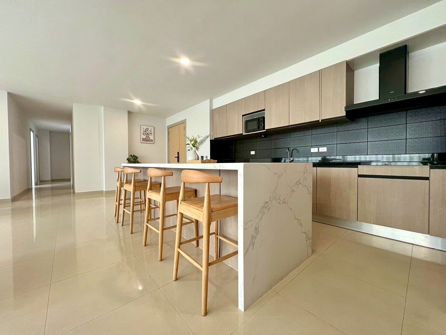 Gardenia_Pattaya_4Bed_Dlc492_2 Modern open kitchen with a marble island and four wooden bar stools, light wood cabinets, and a black tiled backsplash.