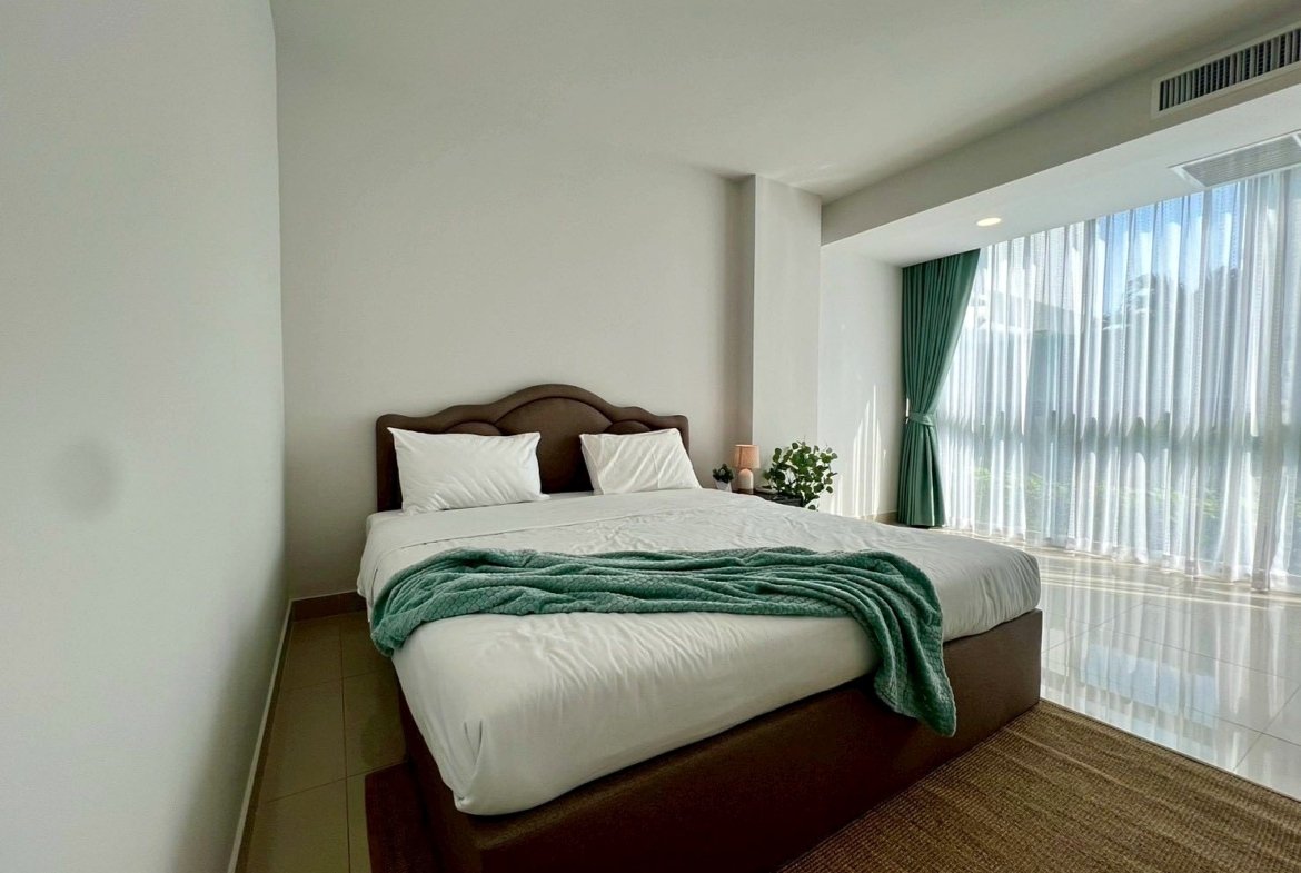 Bright hotel bedroom with white bedding, teal throw, and a large window covered by sheer curtains and green drapes.