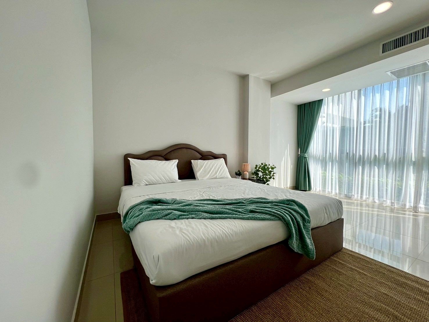 Gardenia_Pattaya_4Bed_Dlc492_20 Bright hotel bedroom with white bedding, teal throw, and a large window covered by sheer curtains and green drapes.