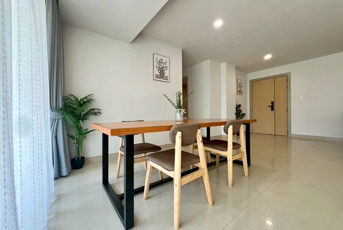 Minimalist dining area: long wooden table with black legs, three wooden chairs, and a potted plant in the corner.