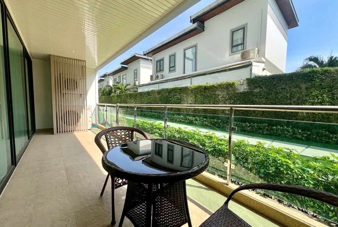 Balcony with glass railing, round glass table and wicker chairs overlooking a green hedge and white modern houses in the background.