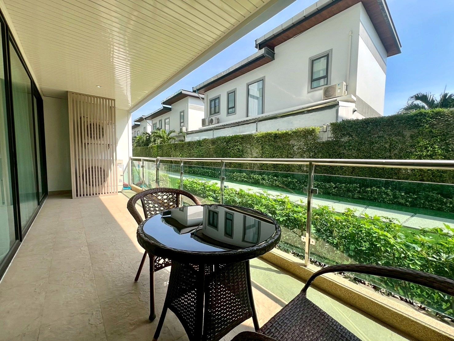 Gardenia_Pattaya_4Bed_Dlc492_28 Balcony with glass railing, round glass table and wicker chairs overlooking a green hedge and white modern houses in the background.
