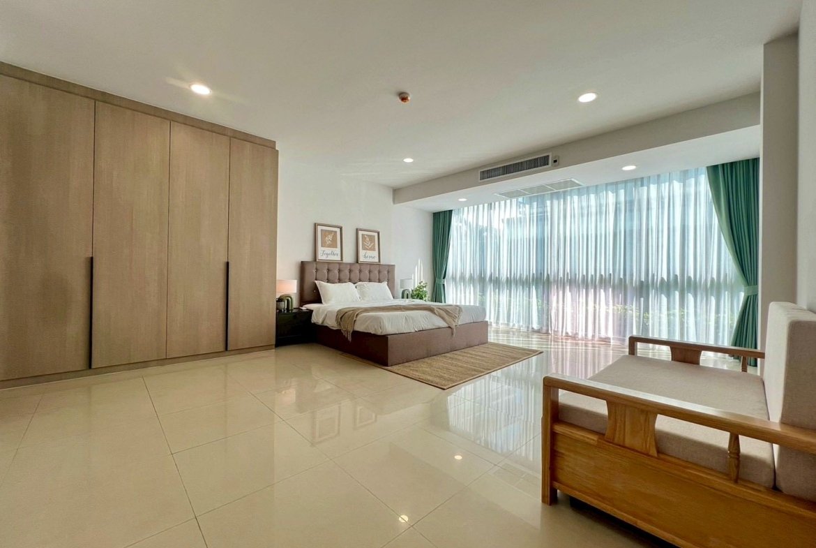 Spacious modern bedroom with a king bed, tufted headboard, and a large wardrobe against the wall near a seating area by tall windows with sheer curtains.