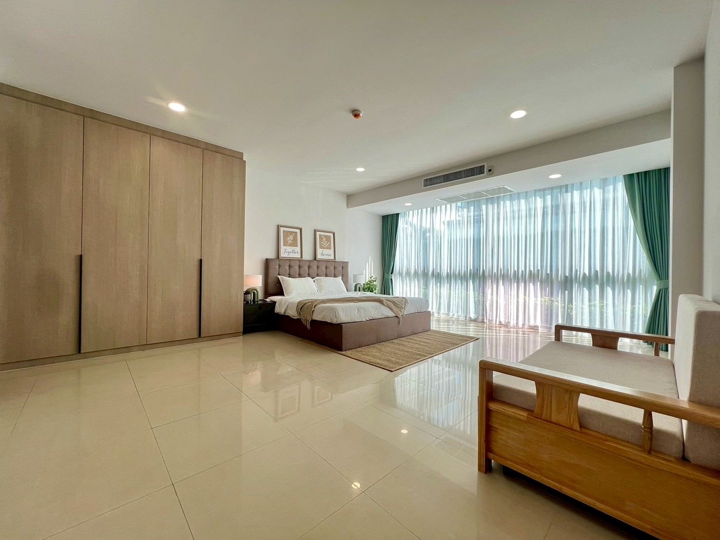 Gardenia_Pattaya_4Bed_Dlc492_4 Spacious modern bedroom with a king bed, tufted headboard, and a large wardrobe against the wall near a seating area by tall windows with sheer curtains.