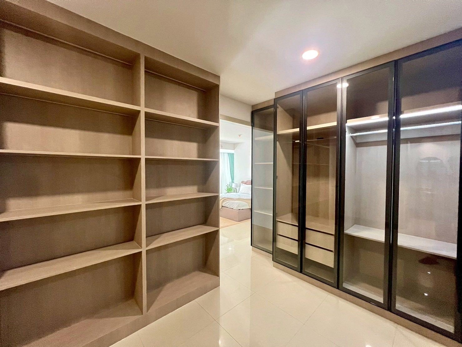 Gardenia_Pattaya_4Bed_Dlc492_8 Walk-in closet with tall built-in wooden shelves on the left and glass-front wardrobes on the right, reflecting a modern, organized space.
