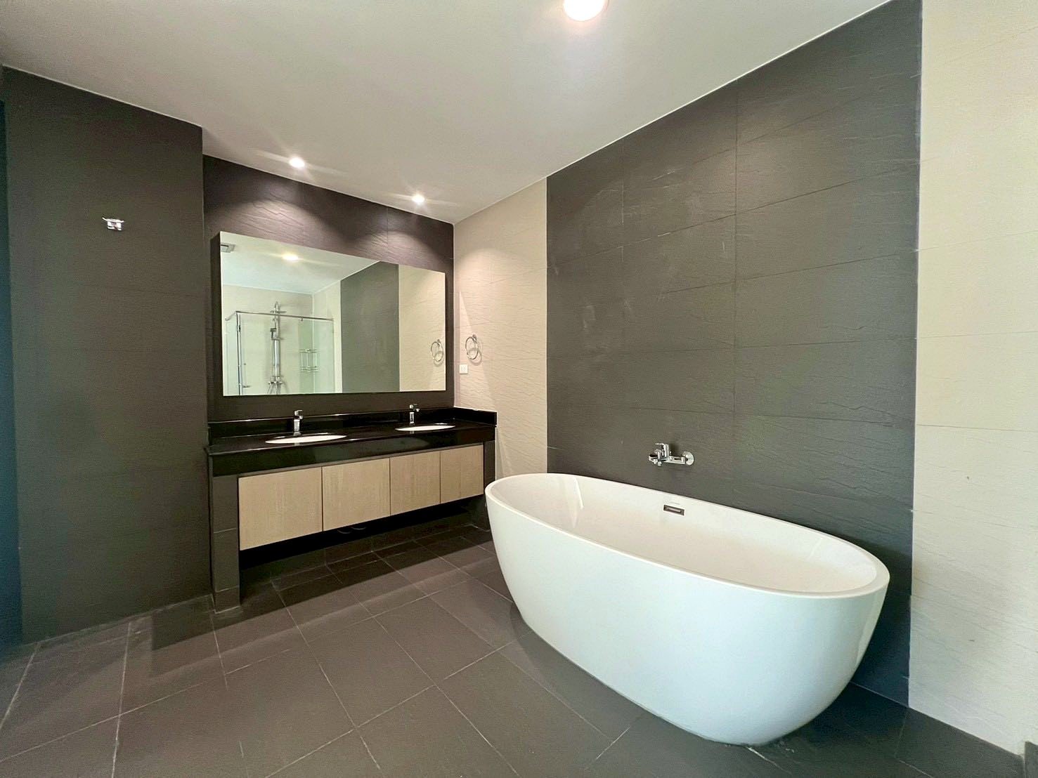 Gardenia_Pattaya_4Bed_Dlc492_9 Contemporary bathroom with a white freestanding oval tub, dark tiled walls, and a double-sink vanity with a large mirror.