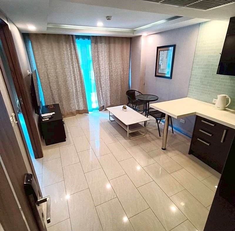 Compact hotel suite living area with kitchenette, coffee table, two chairs, TV stand, and large curtained window.