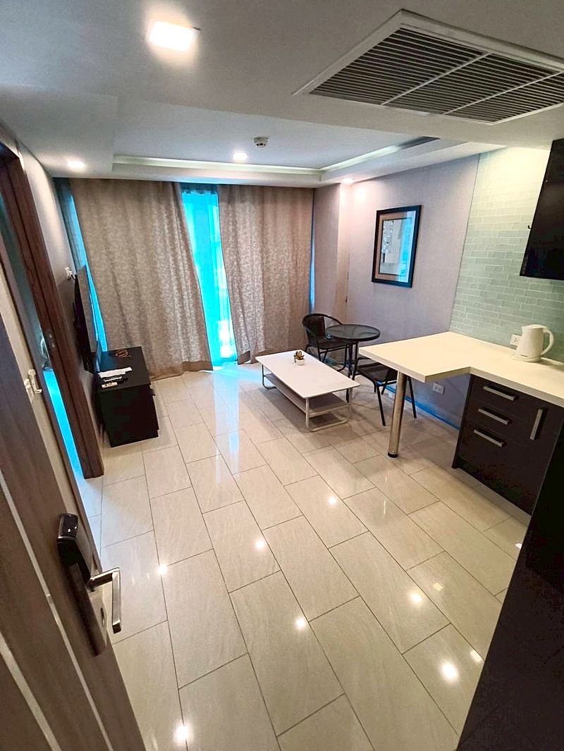 Grand_Avenue_Pattaya_Sale_Dlc641 Compact hotel suite living area with kitchenette, coffee table, two chairs, TV stand, and large curtained window.