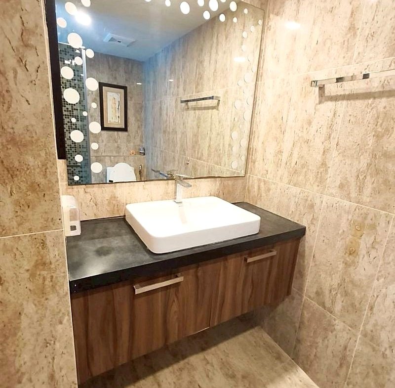 Bathroom vanity with a white rectangular sink on a dark countertop and wooden cabinets beneath, reflected in a large mirror with white polka dots.