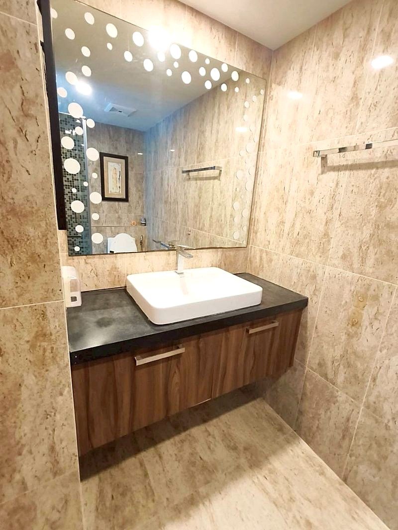 Grand_Avenue_Pattaya_Sale_Dlc641_5 Bathroom vanity with a white rectangular sink on a dark countertop and wooden cabinets beneath, reflected in a large mirror with white polka dots.