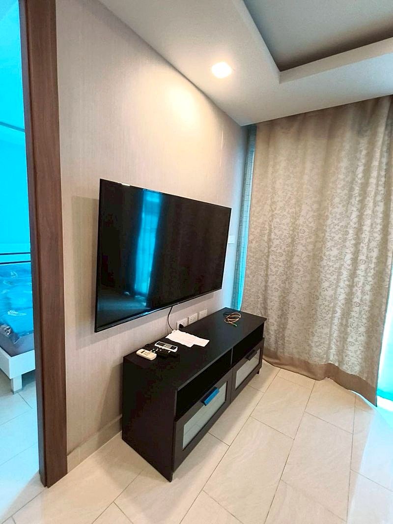 Grand_Avenue_Pattaya_Sale_Dlc641_8 Flat-screen TV mounted on a light wall above a dark TV console with remotes and papers nearby.