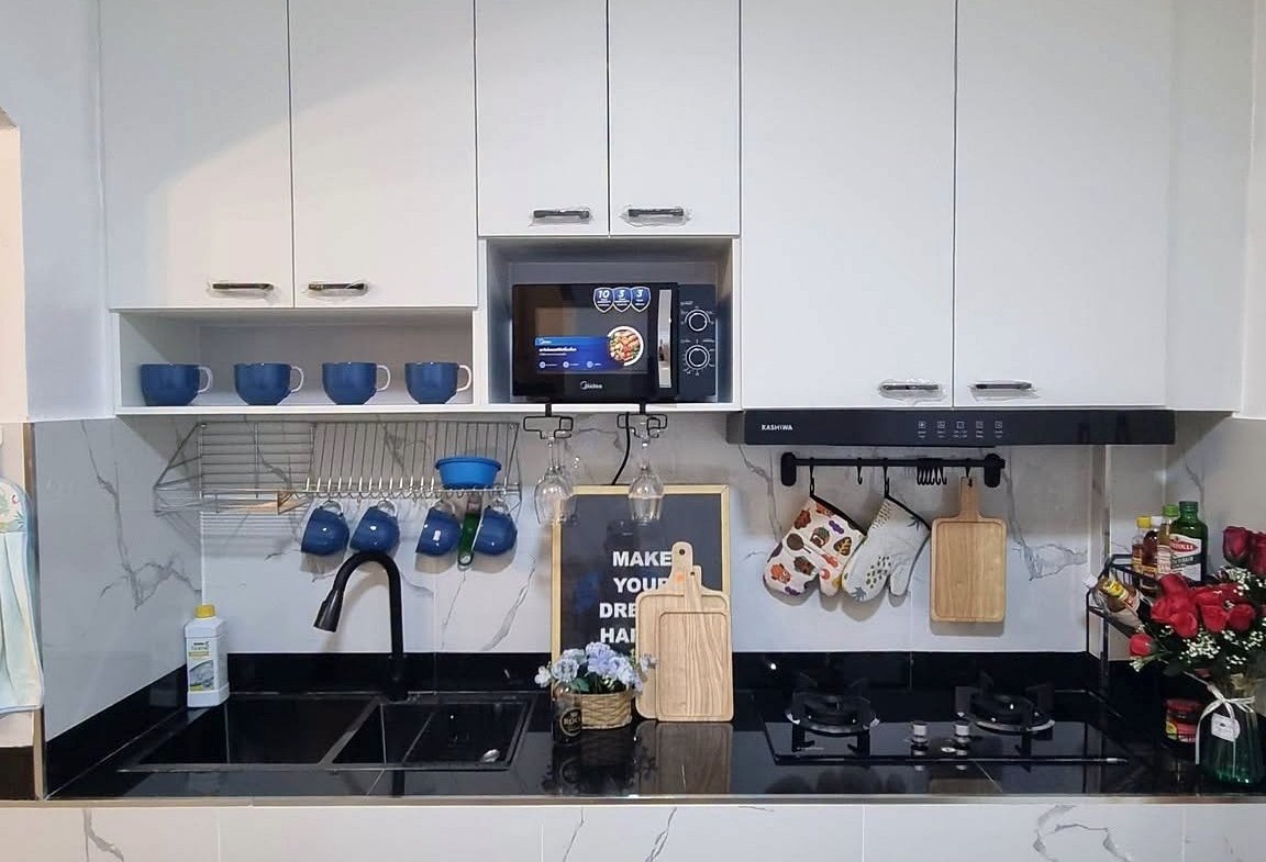White kitchen with upper cabinets, built-in microwave, blue mugs on a rack, black countertop, sink and stove.
