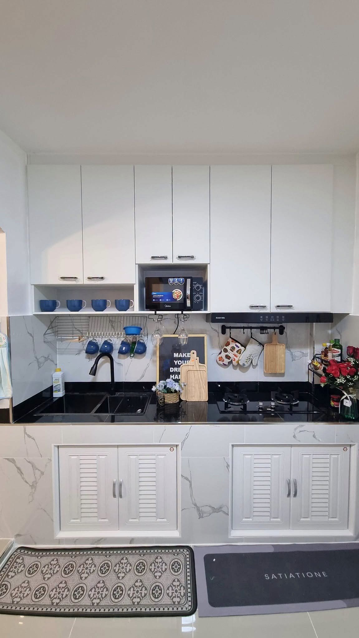 White kitchen with upper cabinets, built-in microwave, blue mugs on a rack, black countertop, sink and stove.