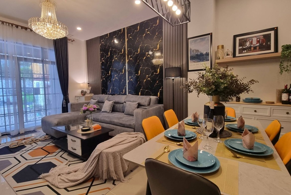 Open-plan living and dining area with a crystal chandelier, gray sectional sofa, marble-pattern feature wall, and a dining table set with turquoise plates and yellow chairs.