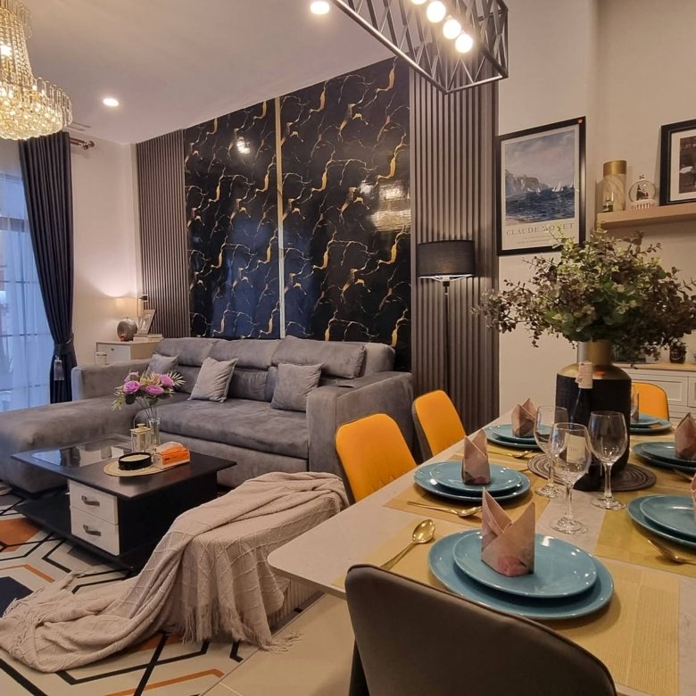 Open plan living and dining area with a crystal chandelier gray sectional sofa marble pattern feature wall and a dining table set with turquoise plates and yellow chairs