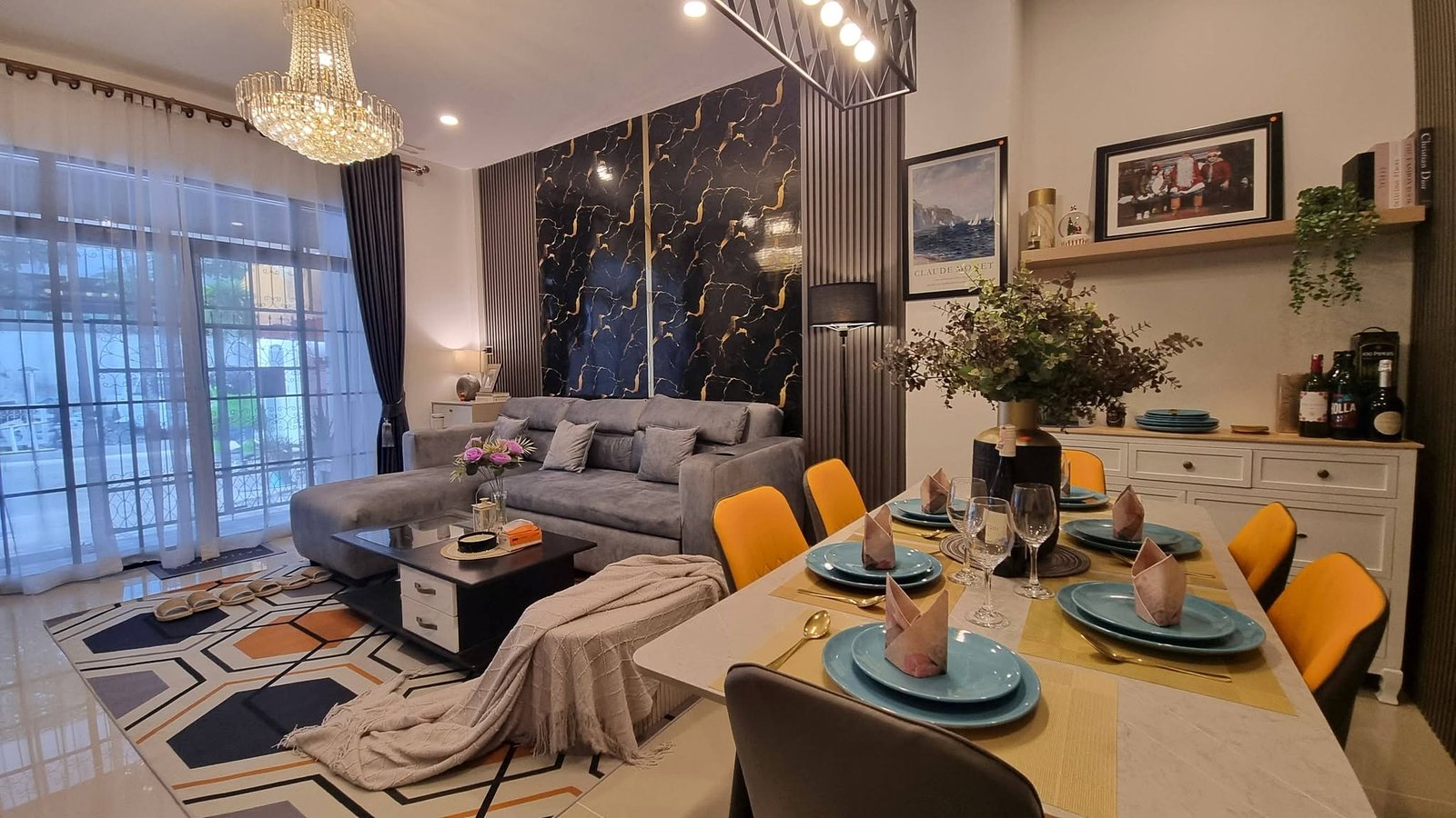 Open-plan living and dining area with a crystal chandelier, gray sectional sofa, marble-pattern feature wall, and a dining table set with turquoise plates and yellow chairs.