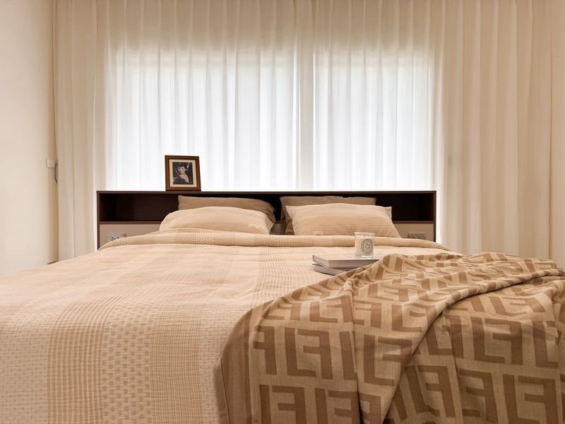 Beige-lyned bed with neutral linens and a geometric throw, framed photo on the headboard shelf, and sheer white curtains in the background.