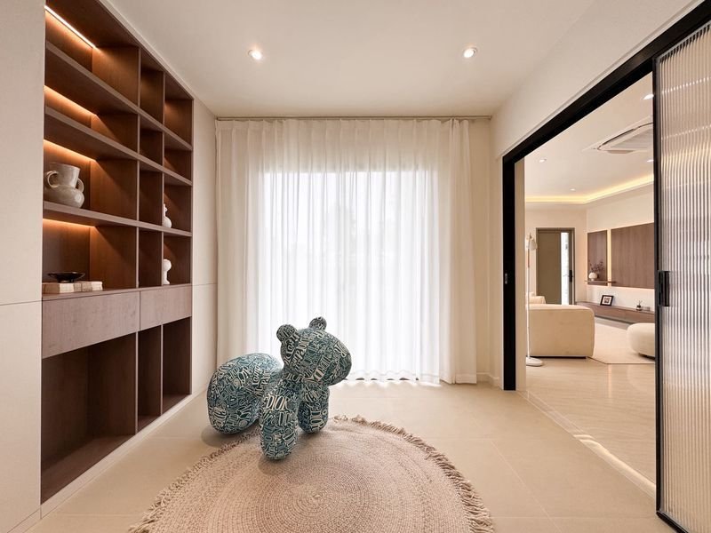 Modern living space with built-in wooden shelves, a blue patterned bear sculpture on a circular rug, sheer white curtains, and a glass sliding door to an adjacent lounge.