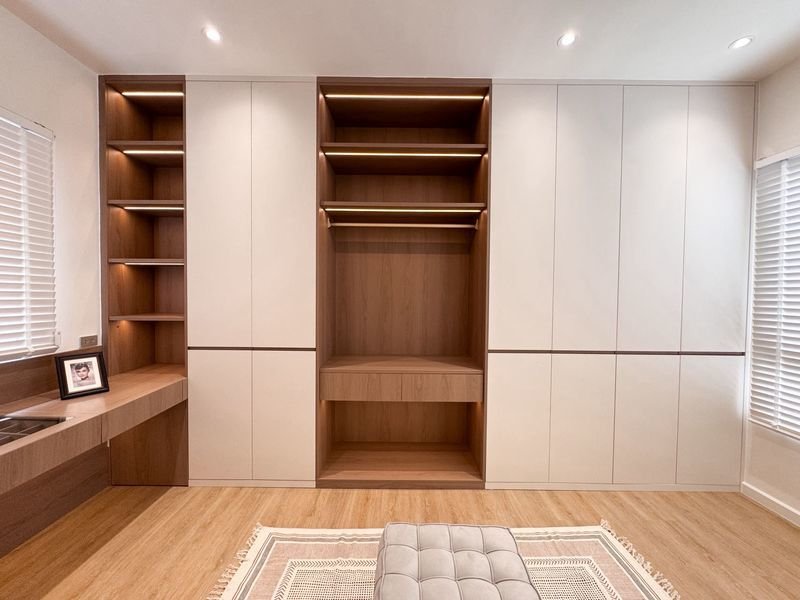 Modern bedroom storage wall with white cabinet doors and open wood shelves surrounding a central open hanging area and lower drawer, warm recessed lighting, and a light wood floor.