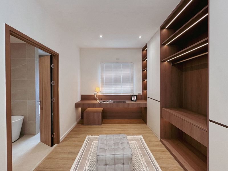 Modern bedroom with built-in wooden shelves and a desk, a tufted ottoman on a rug, and a window with blinds in back.