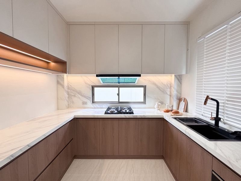 Modern L-shaped kitchen with white upper cabinets, wood base cabinets, marble countertops, gas cooktop under a window, and a black sink with a tall faucet.