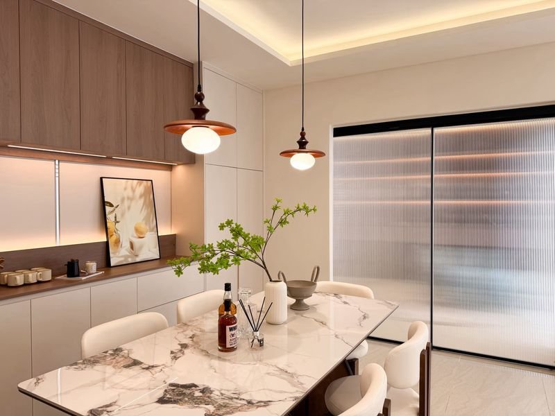 Modern kitchen and dining area with a marble table, bottle and glass, and a potted branch centerpiece under copper pendant lights.