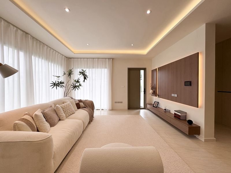 Spacious modern living room with a long beige sofa and many pillows, sheer white curtains, and a plant in a corner.