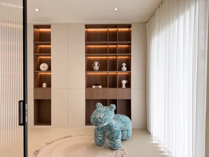 Modern living space with built-in cabinets and open shelving lit by warm LEDs, a blue patterned stuffed bear on a circular rug by sheer white curtains.