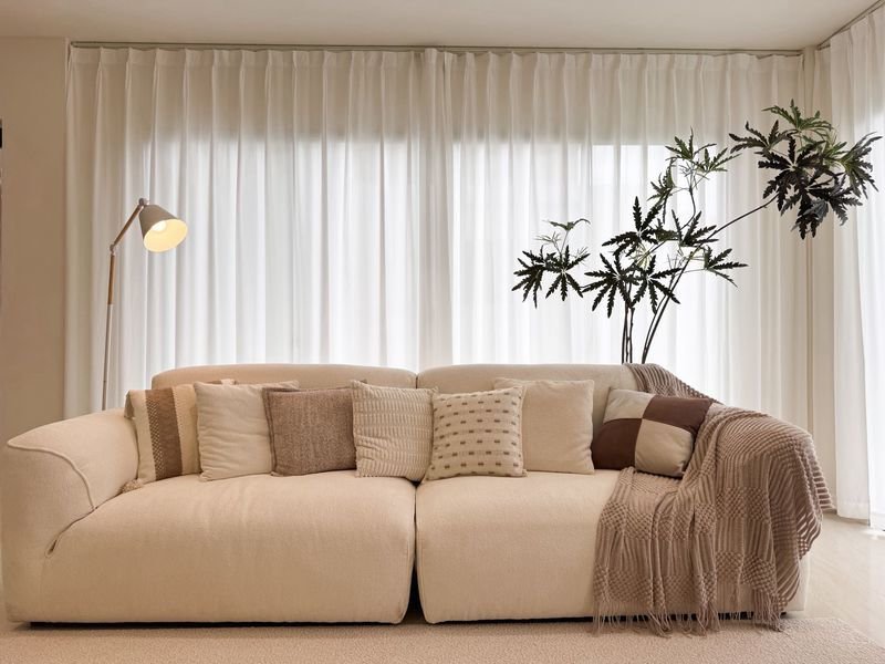 Beige sectional sofa with multiple throw pillows, a floor lamp on the left, sheer white curtains, and a tall plant on the right in a bright living room.