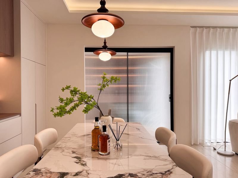 Modern dining area with a marble table, greenery centerpiece, bottles and diffuser, white chairs, and pendant lights.