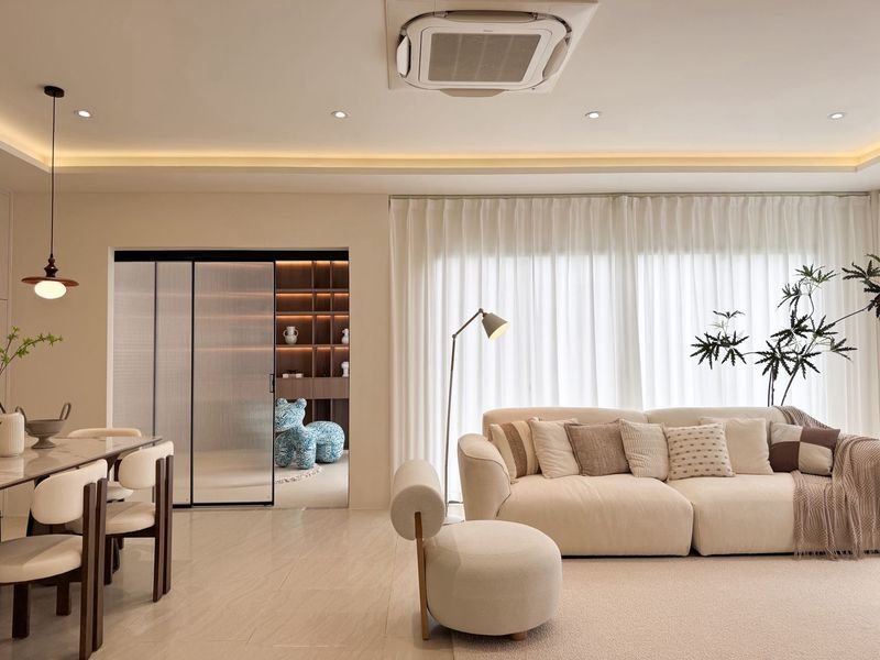 Modern cream living room with a beige sofa, neutral cushions, and a tall floor lamp by a large window with sheer white curtains.