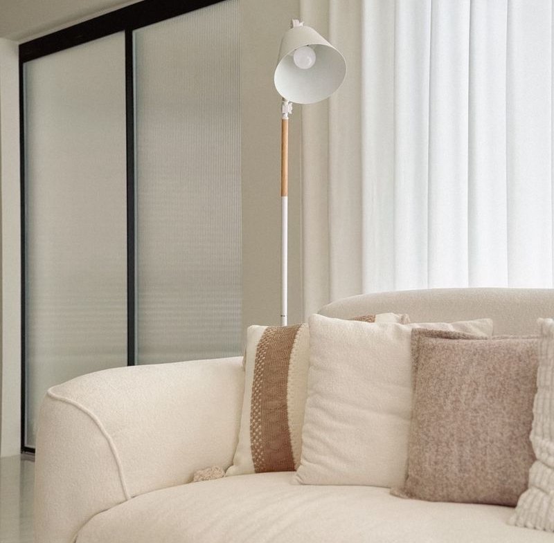 White curved sofa with beige and cream cushions in a bright living room; a white floor lamp stands behind it near sheer curtains.