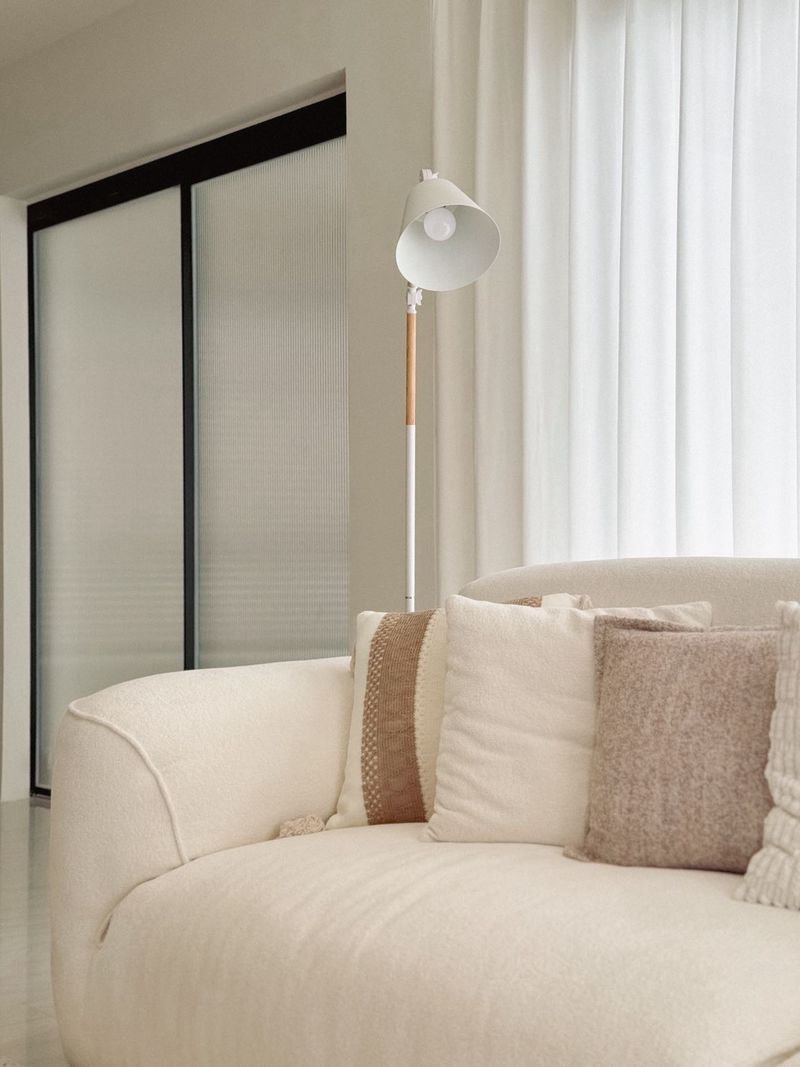 Horizon_By_Patta_10-5mb_Dlh537_9 White curved sofa with beige and cream cushions in a bright living room; a white floor lamp stands behind it near sheer curtains.