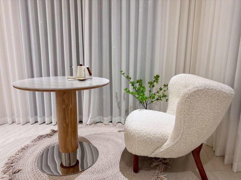 Cozy reading corner with a round wooden table, a white boucle chair, a green plant, and a beige rug in front of white curtains.