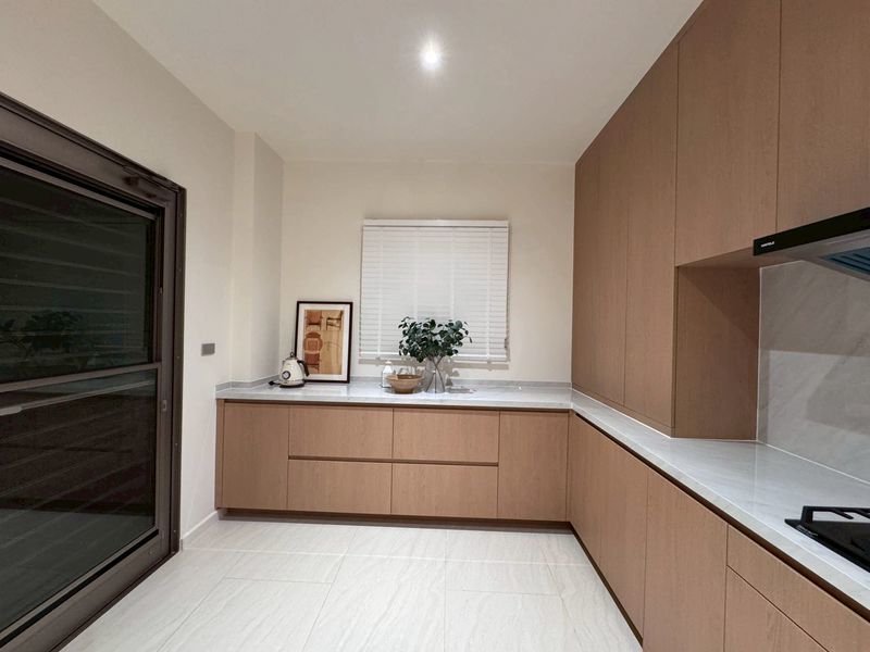 Modern L-shaped kitchen with light wood cabinets, white countertops, a plant on the window sill, and a framed picture on the counter on the left side of the windowed wall.