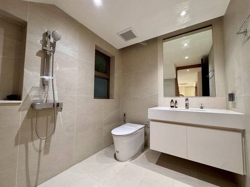 Modern beige-tiled bathroom featuring a wall-mounted handheld shower, a white toilet, and a floating vanity with a large mirror above it. Neutral, minimal design with recessed ceiling lighting.