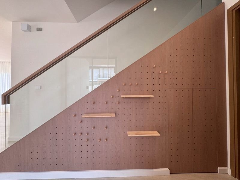 Diagonal wooden pegboard wall with three small beige shelves under a glass staircase railing in a bright modern foyer.