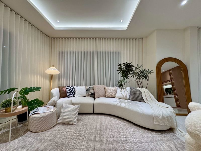 Bright living room with a curved white sofa adorned with patterned cushions, cream rug, and tall plants by sheer curtains.