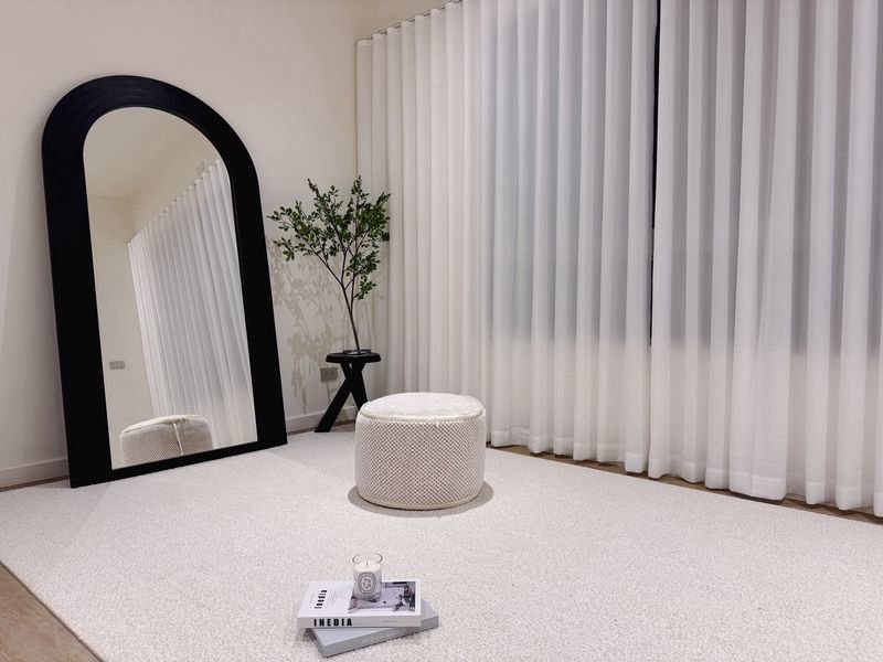 Minimalist room with a tall black arched mirror, beige pouf, plant, and white sheer curtains along the window.