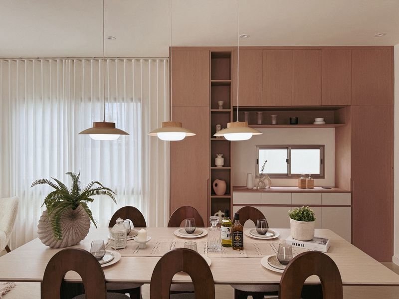 Long wooden dining table set for six with place settings, pendant lights, and a warm wood-toned open kitchen in the background.