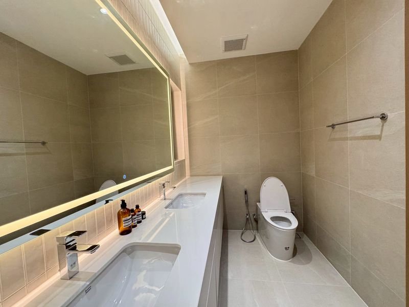 Modern bathroom with a double-sink vanity, backlit mirror, and beige tiled walls surrounding a white toilet.