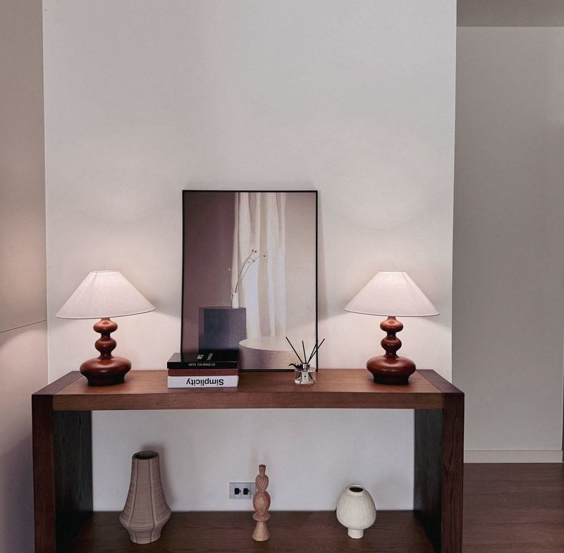 Wood console table with two matching white-shaded lamps flanking a framed image, against a white wall.